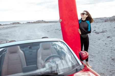 Portrait of a young woman surfer in swimsuit standing with surfboard behind her red car on the rocky coastの写真素材