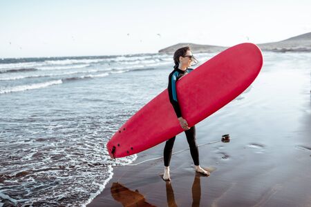Portrait of a young woman surfer in swimsuit standing with red surfboard on the beach. Active lifestyle and surfing conceptの写真素材