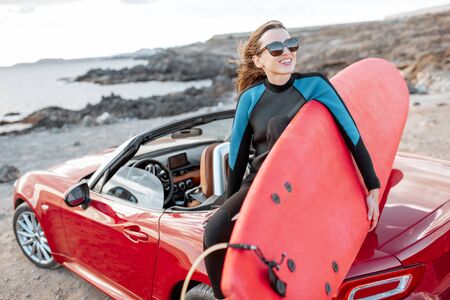 Portrait of a young woman surfer in swimsuit sitting with surfboard on the red cabriolet on the rocky ocean coast. Active lifestyle and surfing conceptの写真素材