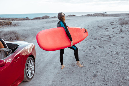Portrait of a young woman surfer in swimsuit standing with surfboard on the dirt road near the...