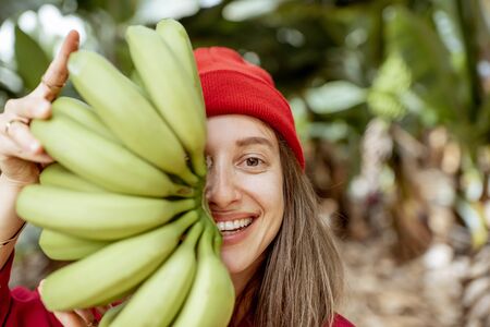 Facial portrait of a cute woman holding a stem with fresh green bananas in front of her face. Healthy eating and wellness conceptの写真素材