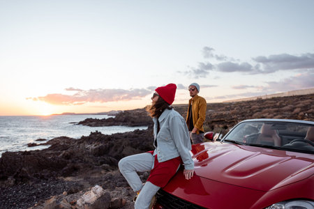 Young joyful couple having fun on the rocky coast while traveling by car on a sunset. Carefree...