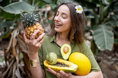 Portrait of a young smiling woman with exotic food outdoors, holding ananas, papaya, melon, avocado and cherymoya fruits. Concept of vegetarianism, healthy eating and wellbeingの写真素材