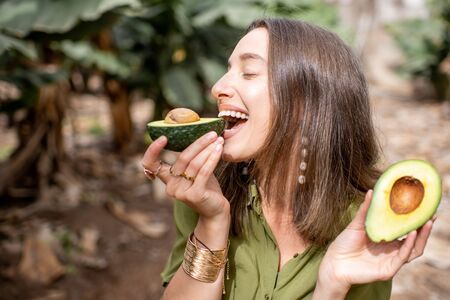 Young smiling woman eating avocado outdoors, standing on the tropical plantation. Concept of vegetarianism, healthy eating, skin care and wellbeingの写真素材