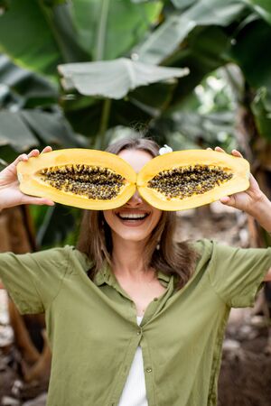 Closeup portrait of a young smiling woman hiding her eyes behind sliced papaya fruit on the green background. Skin care and wellbeing conceptの写真素材