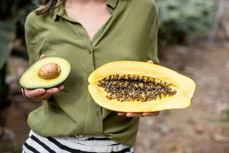 Woman holding sliced papaya fruit and avocado berry outdoors on the tropical plantation, closeup. Concept of vegetarianism, healthy eating and wellbeingの写真素材