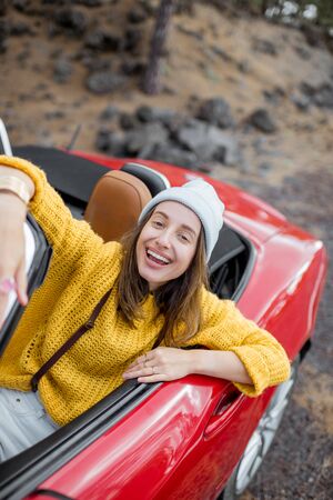 Portrait of a stylish woman in yellow sweater sitting on the drivers seat of convertible sports car on the roadside. Carefree lifestyle and traveling conceptの写真素材