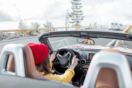 Happy woman dressed in red hat and sweater driving convertible car on the beautiful mountain road, view from the backsideの写真素材