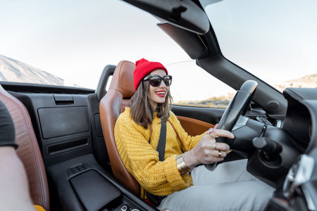 Happy woman in bright hat and sweater driving convertible car while traveling on the desert road on...