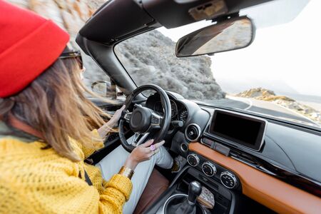 Happy woman in bright hat and sweater driving convertible car while traveling on the desert road on a sunsetの写真素材