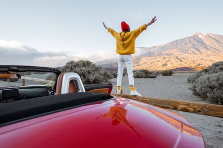 Woman dressed in bright sweater and hat enjoying great mountain landscapes while standing back on the road fence. Traveling by car on Tenerife island, wide view