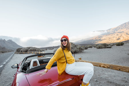 Portrait of a stylish woman dressed in bright sweater and hat enjoying road trip, sitting on the car...