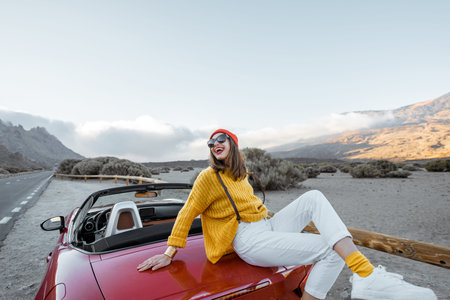 Portrait of a stylish woman dressed in bright sweater and hat enjoying road trip, sitting on the car...