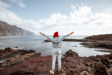 Young woman in red hat enjoying a trip on a rocky ocean coast, standing with raised hands while...