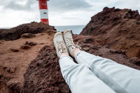 Young traveler resting on the rocky land, traveling volcanic landscapes on the ocean shore near the lighthouse. cropped view focused on the woman's shoesの写真素材