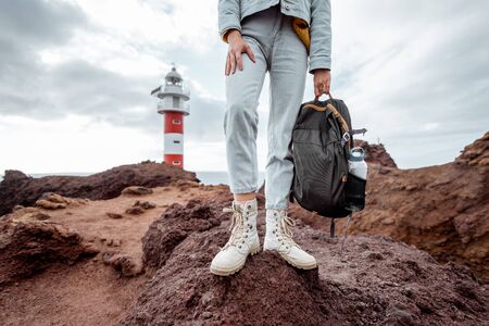 Traveler with backpack standing on the rocky land, traveling volcanic landsacpes near the lighthouse. View on the woman's trekking shoesの写真素材