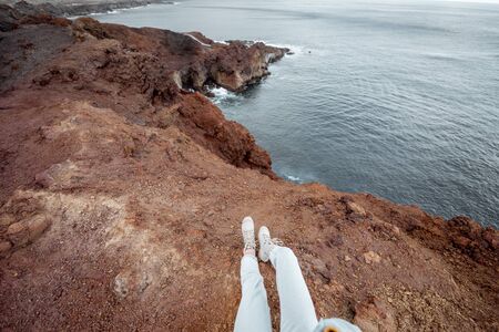 Traveler sitting above the precipice on a rocky coast, traveling on the island, cropped view, focused on women's legs above the oceanの写真素材