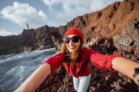 Young stylish woman making selfie portrait while traveling on the rocky ocean coast near the...