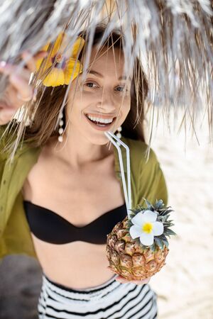 Portrait of a beautiful young smiling woman with exotic cocktail at the beach resort on a sunny day. Concept of exotic summer vacations, beauty and wellnessの写真素材