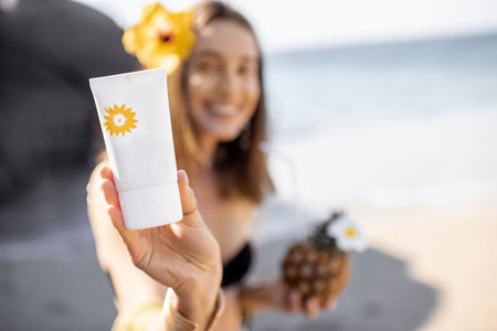Beautiful smiling girl showing tube with sunscreen lotion while resting on the beach, image focused on the tube. Sun protection concept for skinの写真素材