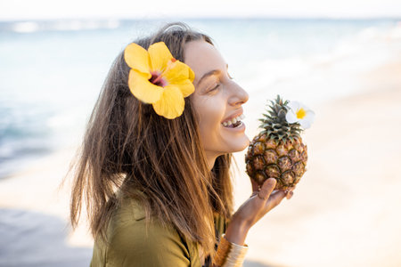 Portrait of a beautiful smiling woman with hair flower and pineapple fruit at the beach on a sunny day. Concept of an exotic summer vacations, beauty and wellnessの写真素材