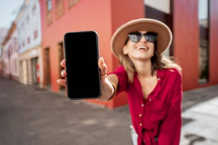 Young stylish woman showing smart phone with blank screen to copy paste while standing on the city street with colorful buildings behind. Image focused on the phoneの写真素材