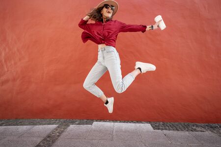Portrait of an active stylish woman dressed in bright shirt with hat and coffee cup jumping on the red wall background outdoors. Carefree lifestyle, coffee and womens fashion conceptの写真素材