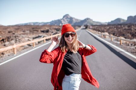 Lifestyle portrait of a young woman stylishly dressed in red walking on the beautiful road in the midst of volcanic valley on a sunny day. Carefree lifestyle and travel conceptの写真素材