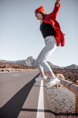 Lifestyle portrait of a young woman stylishly dressed in red jumping on the road, traveling on the...