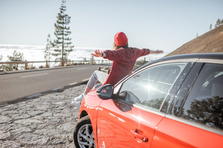 Carefree woman dressed casually sitting on the car hood, enjoying road trip on the mountain road...