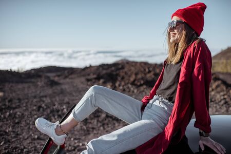 Lifestyle portrait of a stylish woman dressed in red shirt and hat enjoying beautiful sunset above the clouds, traveling by car on the mountainsの写真素材