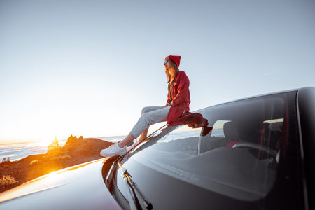 Young woman dressed in red enjoying sunset landscape above the clouds, sitting on the car roof while...