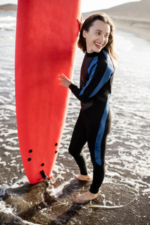 Portrait of a young woman in wetsuit standing with red surfboard on the ocean beach during a sunset....