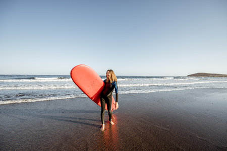 Young surfer in wetsuit walking with red surfboard on the beautiful ocean beach during a sunset,...