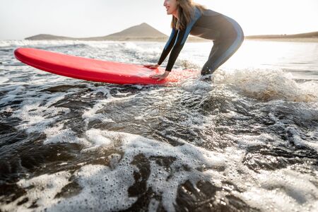 Young surfer in wetsuit getting on surfboard, catching water flow near the beach during a sunset....