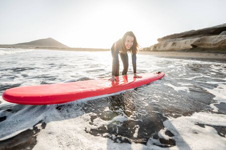 Young surfer in wetsuit getting on surfboard, catching water flow near the beach during a sunset. Water sports and active lifestyle conceptの写真素材