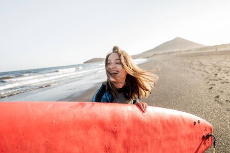 Portrait of a young joyful woman in wetsuit carrying surfboard on the ocean beach during a sunset. Water sport and active lifestyle conceptの写真素材
