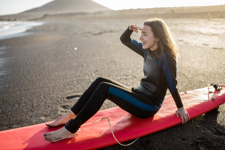 Portrait of a young relaxed surfer in wetsuit sitting with surfboard on the beach. Water sport and...