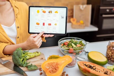 Woman holding digital tablet with launched online shopping market while standing on the kitchen with lots of fresh food on the tableの写真素材