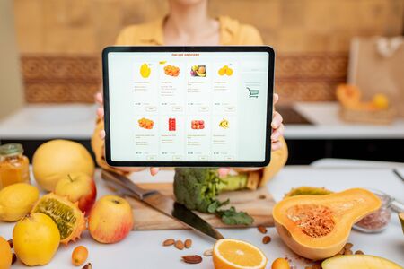 Woman holding digital tablet with launched online shopping market while standing on the kitchen with lots of fresh food on the tableの写真素材