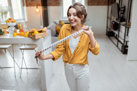 Portrait of a young and cheerful woman with measuring tape at home. Concept of wellbeing and weight...
