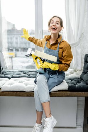 Portrait of a happy young housewife in working clothes washing windows with professional tools at homeの写真素材