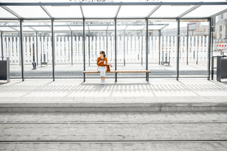 Woman sitting alone at the public transport stop on a sunny day outdoors, general plan on a modern...