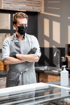 Portrait of a salesman in protective gloves and face mask standing in the shop or cafe. Concept of new social rules for business during a pandemicの写真素材