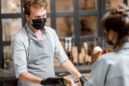 Seller in protective mask and gloves makes contactless payment for a client in the ice cream shop....