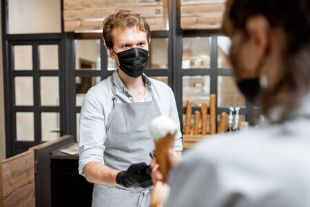 Salesman in protective mask selling ice cream for a young client in the shop. Concept of new social rules for business during a pandemicの写真素材