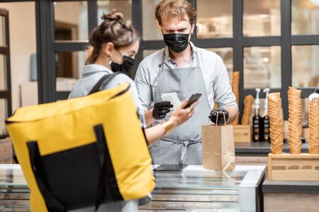 Female courier taking online order for delivery at the counter with salesperson in the small pastry shop or cafe. Concept of a take away food during pandemicの写真素材
