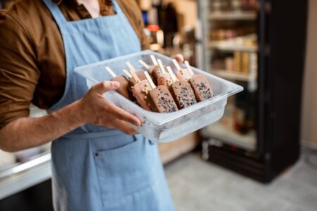 Seller holding a tray with chocolate ice cream on a stick in the shop, close-up on ice cream on a blue apron backgroundの写真素材