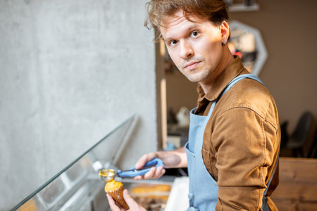 Portrait of a cheerful salesman putting ice cream into the waffle cone at the counter of a shop
