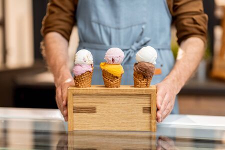 Seller putting a stand with three yummy ice creams in waffle cones with different flavor on the counter of a shop, close-upの写真素材